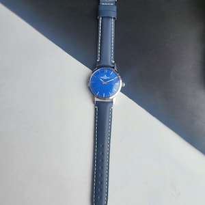 May include: A blue wristwatch with a silver case and a matching blue leather strap. The watch face is a vibrant blue with silver hands and markers. The strap is stitched with white thread.