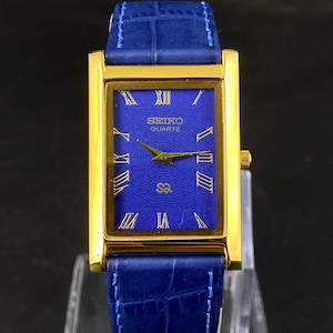 May include: A gold-toned rectangular Seiko Quartz watch with a blue face and blue leather band. The watch face features gold Roman numerals and hands. The watch is displayed on a clear stand.