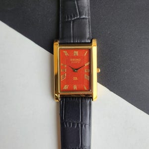 Puede incluir: Reloj Seiko Quartz dorado con esfera rectangular roja y agujas negras. El reloj presenta números romanos y una correa de cuero negro. El fondo es blanco y negro.