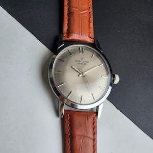 May include: A vintage Breitling Geneve watch with a silver face and a brown leather strap. The watch features silver hands and markers, and a small seconds dial. The case is stainless steel.
