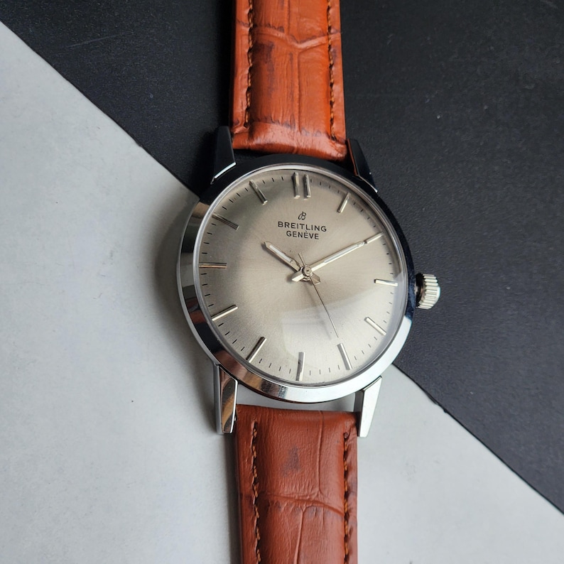 May include: A vintage Breitling Geneve watch with a silver face and a brown leather strap. The watch features silver hands and markers, and a small seconds dial. The case is stainless steel.