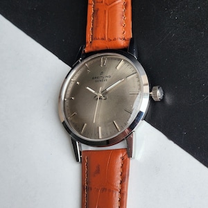 May include: A silver Breitling Gen&egrave;ve wristwatch with a brown leather strap. The watch features a silver face with silver hands and markers. The strap is a textured orange-brown colour.