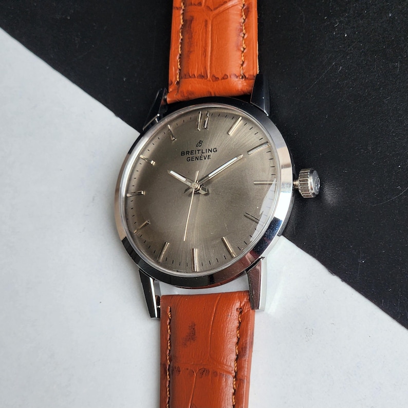 May include: A silver Breitling Gen&egrave;ve wristwatch with a brown leather strap. The watch features a silver face with silver hands and markers. The strap is a textured orange-brown colour.
