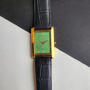 May include: A rectangular gold-toned wristwatch with a green face and black leather band. The watch face features Roman numerals and a textured design. The band has a crocodile pattern. The watch is displayed against a black and white background.