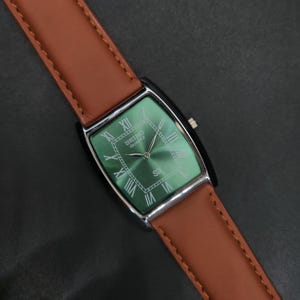 May include: A rectangular Seiko Quartz wristwatch featuring a metallic green dial with Roman numeral hour markers, silver-tone hands, and a brown leather strap with visible stitching. The watch case is silver-tone with a crown on the right side.