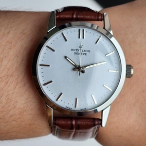 Op de afbeelding: Een zilveren Breitling Gen&egrave;ve horloge met een witte wijzerplaat en een bruine leren band. Het horloge heeft zilveren wijzers en uurmarkeringen. Het horloge zit om een pols.