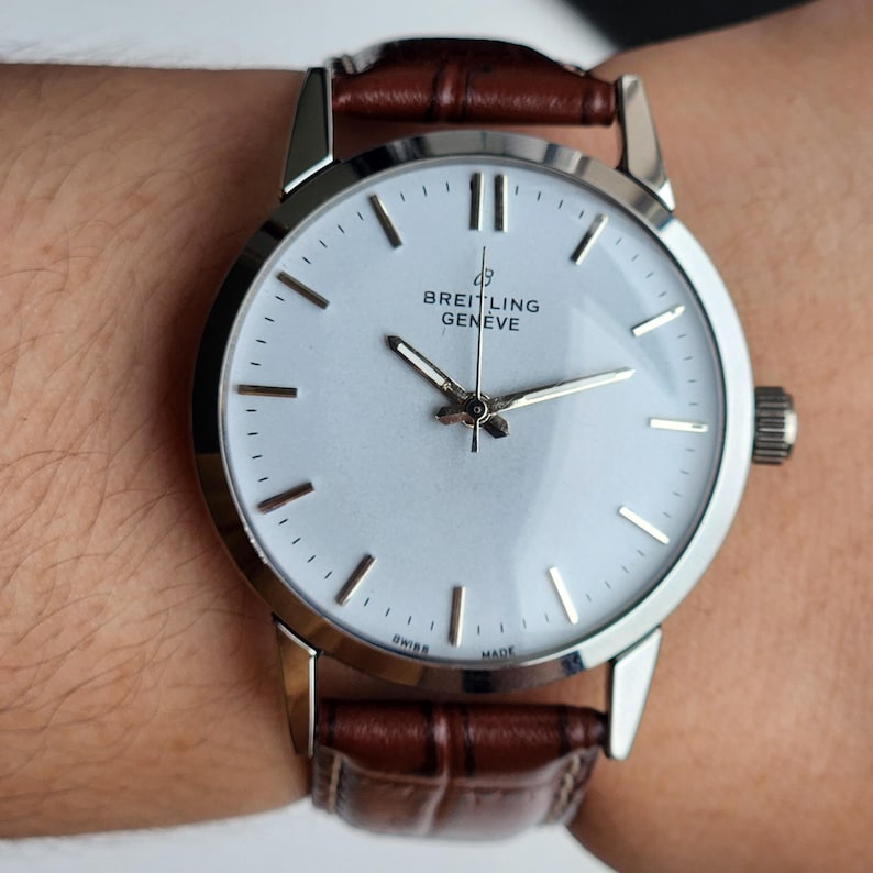 Op de afbeelding: Een zilveren Breitling Gen&egrave;ve horloge met een witte wijzerplaat en een bruine leren band. Het horloge heeft zilveren wijzers en uurmarkeringen. Het horloge zit om een pols.