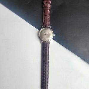 May include: A silver-toned wristwatch with a brown leather band. The watch face is a light silver color, and the band has a textured, crocodile-like pattern. The watch is displayed against a black and white background.