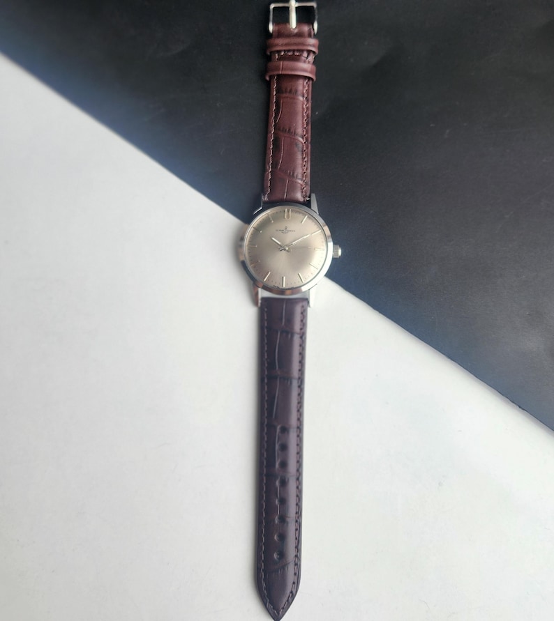 May include: A silver-toned wristwatch with a brown leather band. The watch face is a light silver color, and the band has a textured, crocodile-like pattern. The watch is displayed against a black and white background.
