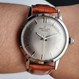 May include: A silver Breitling Gen&egrave;ve wristwatch with a brown leather strap. The watch has a silver face with silver hands and markers. The watch is attached to a brown leather strap.