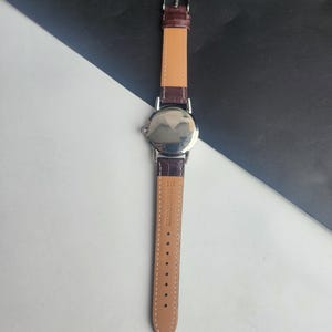 May include: A silver-toned wristwatch with a brown leather band. The watch face is reflective, and the band is secured with a silver buckle. The watch band is made of genuine leather.