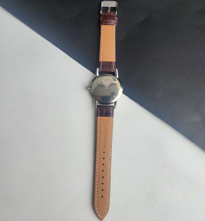 May include: A silver-toned wristwatch with a brown leather band. The watch face is reflective, and the band is secured with a silver buckle. The watch band is made of genuine leather.