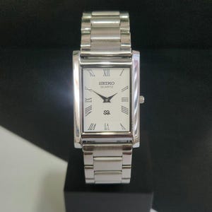 May include: A rectangular Seiko Quartz watch with a silver band and white face. The watch face features black Roman numerals, black hands, and the Seiko Quartz logo. The watch is displayed on a black surface.