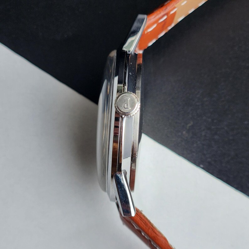 May include: A side view of a silver wristwatch with a brown leather strap. The watch face is round, and the strap is stitched with white thread. The watch has a silver crown with the letter "P" on it.
