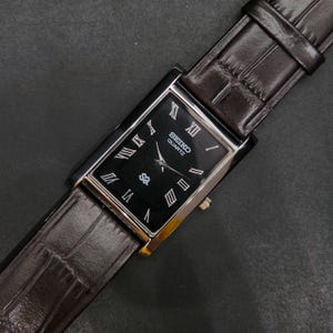 Può includere: Un orologio da polso rettangolare con quadrante nero, indici a numeri romani color argento e lancette argentate. L'orologio presenta la scritta Seiko Quartz sul quadrante e un cinturino in pelle marrone scuro con motivo testurizzato.