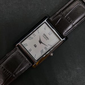 May include: A rectangular Seiko Quartz wristwatch featuring a white dial with black Roman numeral hour markers, black hands, and a logo. The watch is attached to a dark brown leather strap with a textured, embossed pattern and a silver-tone case.