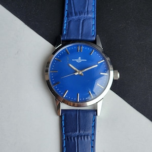 Puede incluir: Un reloj de pulsera con esfera azul vibrante y caja plateada. El reloj presenta una correa de cuero azul con un patr&oacute;n de cocodrilo. La esfera del reloj muestra el nombre de la marca "ULYSSE NARDIN" en texto blanco.