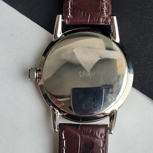 May include: A silver-toned wristwatch with a brown leather band. The watch face is round and reflective, with a small crown on the side. The band is a dark brown with a textured, crocodile-like pattern.