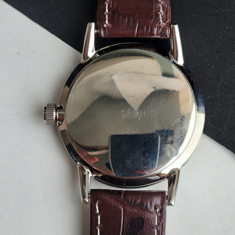 May include: A silver-toned wristwatch with a brown leather band. The watch face is round and reflective, with a small crown on the side. The band is a dark brown with a textured, crocodile-like pattern.