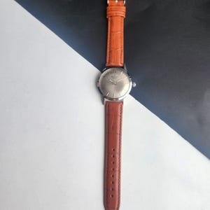 May include: A silver wristwatch with a brown leather strap. The watch face is silver with simple markers and hands. The strap is a reddish-brown colour with a textured, crocodile-style pattern.
