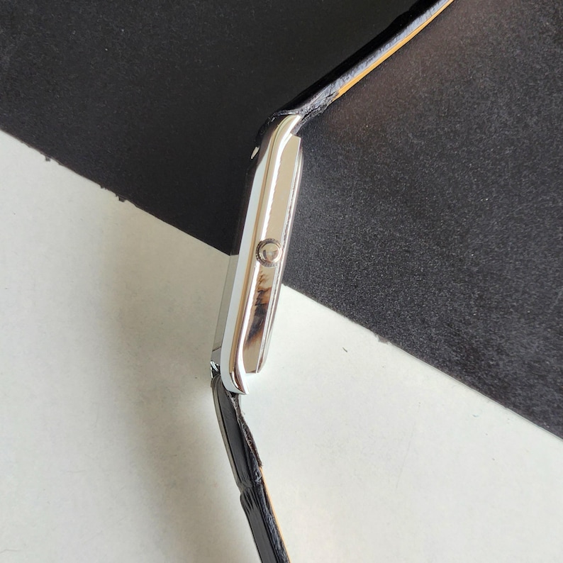 Pode incluir: Um rel&oacute;gio de pulso retangular prateado com uma pulseira de couro preta. O mostrador do rel&oacute;gio &eacute; de cor clara e a pulseira parece ser de couro genu&iacute;no. O rel&oacute;gio tem um design cl&aacute;ssico.