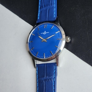Puede incluir: Un reloj de pulsera vintage con una esfera azul vibrante y una correa de cuero azul a juego. El reloj presenta manecillas e indicadores de hora plateados, y la marca "ULYSSE NARDIN" es visible. La caja del reloj es plateada.