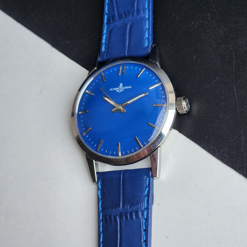 Puede incluir: Un reloj de pulsera vintage con una esfera azul vibrante y una correa de cuero azul a juego. El reloj presenta manecillas e indicadores de hora plateados, y la marca "ULYSSE NARDIN" es visible. La caja del reloj es plateada.