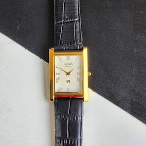 Puede incluir: Un reloj Seiko Quartz rectangular dorado con esfera blanca y correa de cuero negro. La esfera del reloj presenta números romanos y el logotipo de Seiko Quartz. La correa negra tiene un patrón texturizado similar al de un cocodrilo.