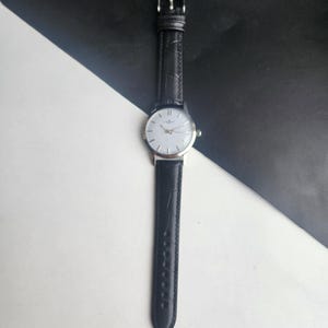 May include: A silver-toned wristwatch with a white face and black leather band. The watch has a classic design with a simple dial and a textured band. The watch is displayed against a black and white background.