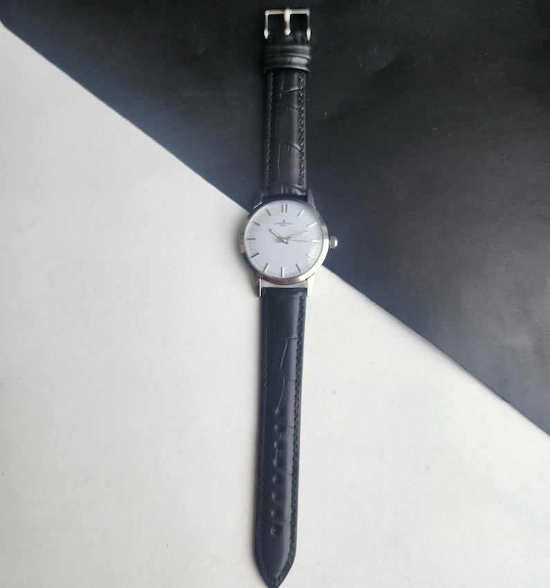 May include: A silver-toned wristwatch with a white face and black leather band. The watch has a classic design with a simple dial and a textured band. The watch is displayed against a black and white background.