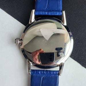 Puede incluir: Un reloj de pulsera plateado con una correa de cuero azul. La esfera del reloj es muy reflectante, mostrando un reflejo distorsionado. La correa es de un azul vibrante y el reloj tiene una peque&ntilde;a corona texturizada.