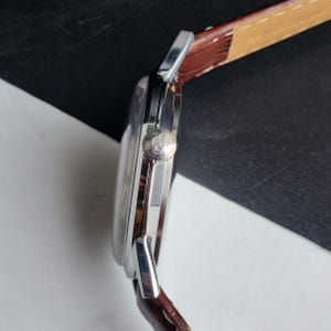 Op de afbeelding: Zijaanzicht van een zilveren polshorloge met een bruine leren band. Het horloge heeft een ronde wijzerplaat en een zilveren kroon. De band is gestikt met witte draad. Het horloge staat tegen een zwart-witte achtergrond.