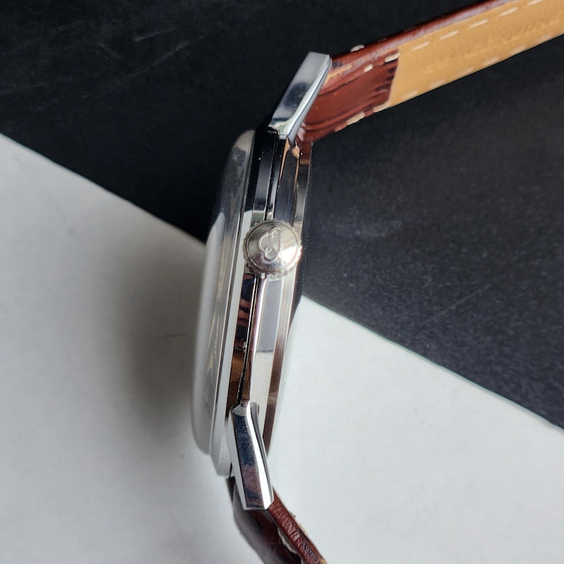 Op de afbeelding: Zijaanzicht van een zilveren polshorloge met een bruine leren band. Het horloge heeft een ronde wijzerplaat en een zilveren kroon. De band is gestikt met witte draad. Het horloge staat tegen een zwart-witte achtergrond.