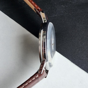 Op de afbeelding: Een zilverkleurig polshorloge met een bruine leren band. De wijzerplaat van het horloge is licht van kleur en de band is gestikt. Het horloge wordt van opzij bekeken.