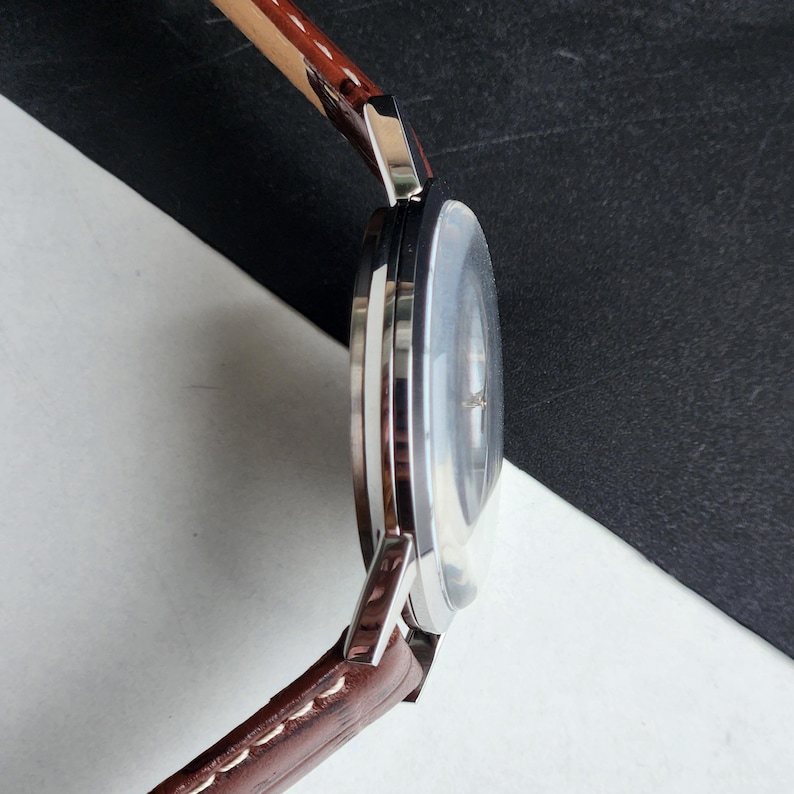 Op de afbeelding: Een zilverkleurig polshorloge met een bruine leren band. De wijzerplaat van het horloge is licht van kleur en de band is gestikt. Het horloge wordt van opzij bekeken.
