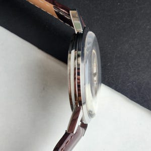 May include: A silver-toned wristwatch with a brown leather band. The watch face is clear, and the band is attached to the watch case with silver-toned metal. The watch is angled, showing the side profile and the band's stitching.