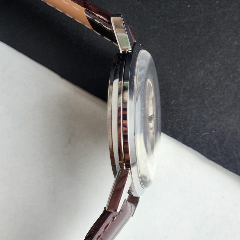 May include: A silver-toned wristwatch with a brown leather band. The watch face is clear, and the band is attached to the watch case with silver-toned metal. The watch is angled, showing the side profile and the band's stitching.