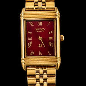 Op de afbeelding: Een rechthoekig goudkleurig Seiko Quartz horloge met een dieprode wijzerplaat en gouden Romeinse cijfers als uurmarkeringen. Het horloge heeft een bijpassende goudkleurige metalen schakelarmband en een kroon aan de rechterkant.