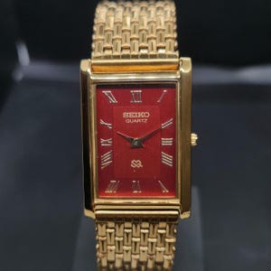 Op de afbeelding: Een goudkleurig rechthoekig Seiko Quartz horloge met een dieprode wijzerplaat voorzien van een subtiel golvend patroon, gouden Romeinse cijfers, gouden wijzers en een gestructureerde goudkleurige metalen schakelarmband met het Seiko logo.