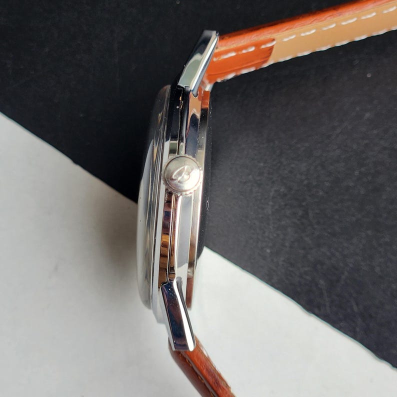 May include: A silver wristwatch with a brown leather strap, viewed from the side. The watch face is black, and the strap is stitched with white thread. The watch has a silver crown with a logo.