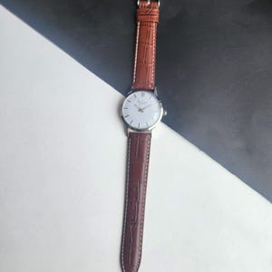 Op de afbeelding: Een zilveren polshorloge met een witte wijzerplaat en bruine leren band. Het horloge heeft een klassiek ontwerp met een ronde wijzerplaat en een leren band met textuur. De band is ongeveer 2 cm breed.
