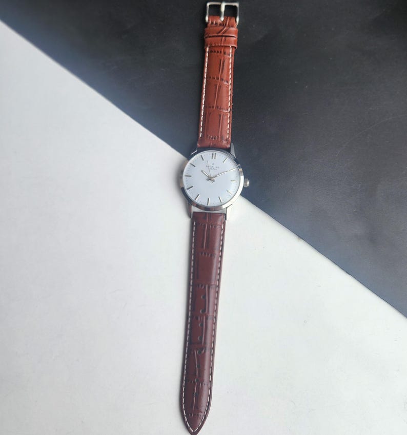 Op de afbeelding: Een zilveren polshorloge met een witte wijzerplaat en bruine leren band. Het horloge heeft een klassiek ontwerp met een ronde wijzerplaat en een leren band met textuur. De band is ongeveer 2 cm breed.