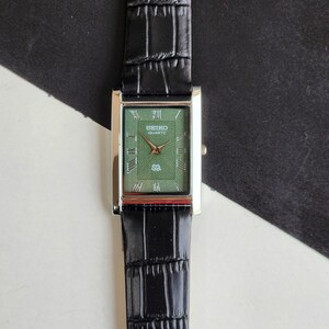 Pode incluir: Um rel&oacute;gio Seiko Quartz retangular com mostrador verde e ponteiros dourados. O rel&oacute;gio tem uma caixa prateada e uma pulseira de couro preta com padr&atilde;o de crocodilo. O mostrador exibe algarismos romanos e o logotipo Seiko Quartz.