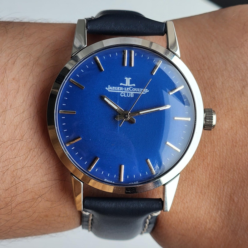 May include: A wristwatch with a blue face and silver case. The watch displays the text "JAEGER-LECOULTRE CLUB" and has a black leather strap. The watch hands and hour markers are silver.