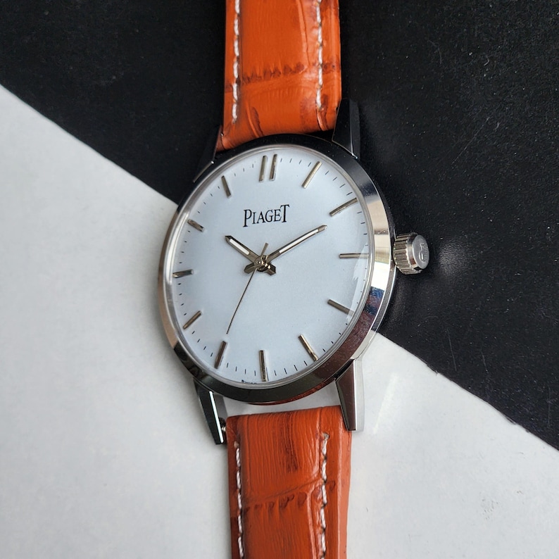 May include: A silver-toned Piaget wristwatch with a white face and black hour markers. The watch has a leather band in a vibrant orange color. The watch face displays the brand name "Piaget".