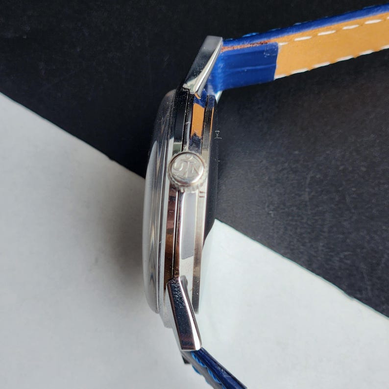 Puede incluir: Un reloj de pulsera plateado con una correa de cuero azul y una parte inferior de color canela. La esfera del reloj es redonda y la correa est&aacute; sujeta con metal plateado. El reloj est&aacute; sobre un fondo blanco y negro.