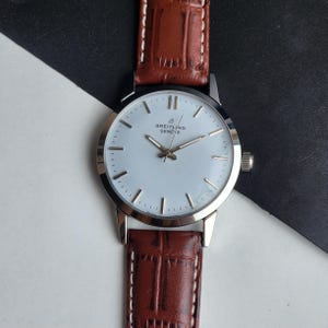 Op de afbeelding: Een Breitling Geneve polshorloge met een lichtblauwe wijzerplaat, zilveren wijzers en een bruine leren band. Het horloge heeft een zilveren kast en een klassiek ontwerp. De band is ongeveer 20 cm lang.