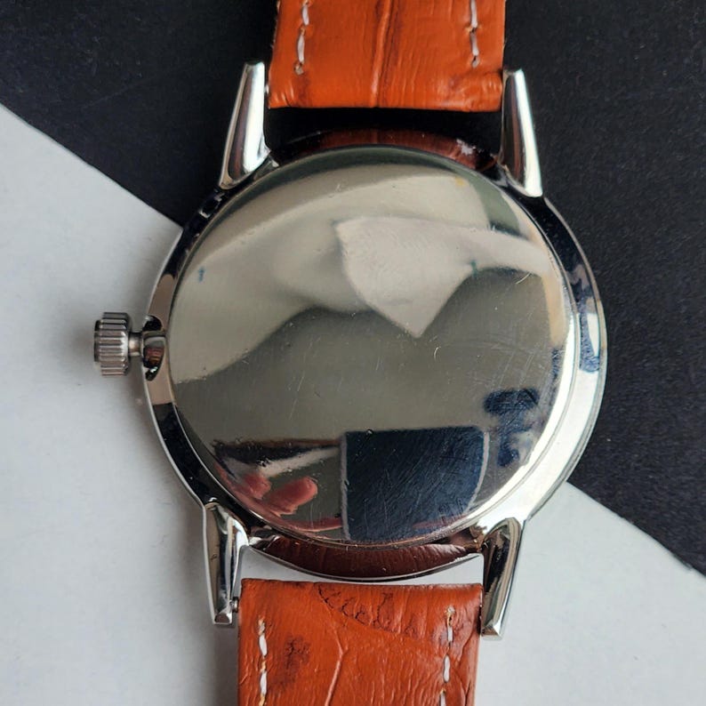 May include: Close-up of the back of a silver wristwatch with a polished, reflective surface. The watch has a brown leather strap with a textured pattern. The watch's crown is visible on the left side.