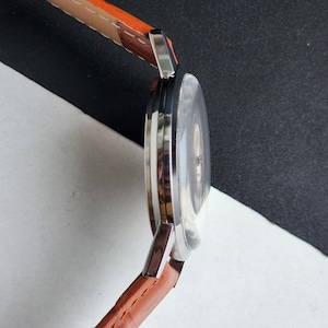 May include: A silver wristwatch with a brown leather strap. The watch face is round and has a silver bezel. The strap is made of brown leather with visible stitching.