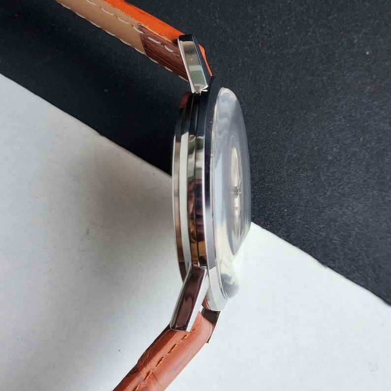 May include: A silver wristwatch with a brown leather strap. The watch face is round and has a silver bezel. The strap is made of brown leather with visible stitching.